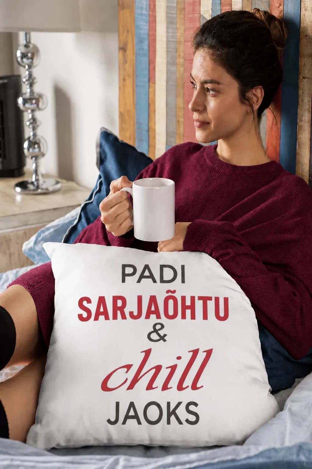 Naine teetassiga diivanil, süles dekoratiivpadi punase ja musta kirjaga „Padi sarjaõhtu & chill jaoks“